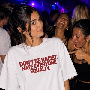Può includere: Una t-shirt bianca a maniche corte con il testo "DON'T BE RACIST. HATE EVERYONE EQUALLY." stampato in lettere maiuscole rosso scuro sul petto. Il capo presenta un girocollo e una vestibilità ampia e rilassata.