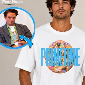 Puede incluir: Una camiseta blanca de manga corta con un gráfico circular de una galleta con chispas de chocolate y la palabra PRIMETIME impresa en letras mayúsculas de color azul claro en el centro del pecho de la prenda.