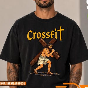 Könnte beinhalten: Ein schwarzes T-Shirt mit kurzen Ärmeln und einem Grafikdruck einer Figur, die ein großes Holzkreuz trägt. Über der Grafik ist das Wort Crossfit in fetter gelber Schrift gedruckt. Das Design zeigt zudem eine Dornenkrone und Sandalen.