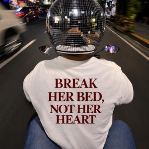 Può includere: Una t-shirt bianca con la scritta rosso scuro "BREAK HER BED, NOT HER HEART" stampata sul retro, indossata con jeans blu e un casco sferico ricoperto di piccole tessere a specchio argentate che creano un effetto palla da discoteca.