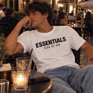 Può includere: Una t-shirt bianca a maniche corte con la scritta nera ESSENTIALS FEAR OF JOB stampata sul petto. Il capo è abbinato a jeans in denim azzurro, creando un look casual. La t-shirt presenta un girocollo standard e una vestibilità rilassata.