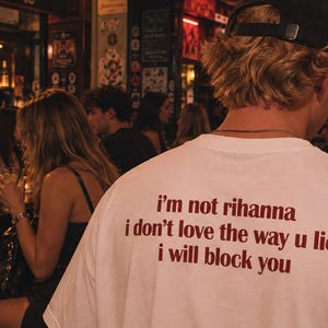 Può includere: Una t-shirt bianca a maniche corte con testo rosso scuro stampato sul retro che recita: i'm not rihanna i don't love the way u lie i will block you. La maglietta è indossata da una persona con capelli biondi e un berretto nero.