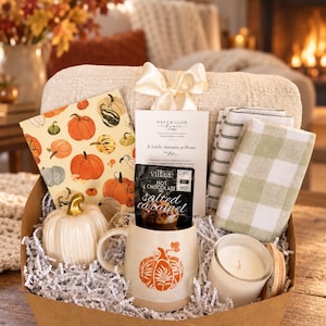 May include: A gift basket containing a pumpkin-patterned notebook, a Nest & Glow card, two checkered napkins, a white ceramic pumpkin, a mug with an orange pumpkin graphic, a packet of salted caramel hot chocolate, and a white candle in a glass jar.