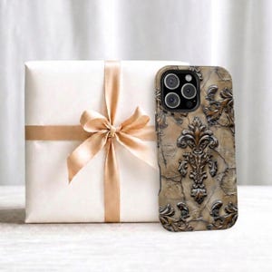 Può includere: Una custodia per smartphone con una superficie testurizzata che ricorda la pietra crepata in tonalità beige e marrone, decorata con un emblema fleur-de-lis in rilievo marrone scuro e motivi a foglia, posizionata accanto a una scatola regalo bianca.