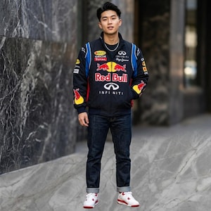 May include: A person wearing a black Red Bull racing jacket with blue and yellow accents, dark blue jeans, and white sneakers with red details. The jacket features the Red Bull logo and Infiniti branding.