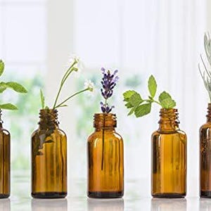 May include: Five amber glass bottles with droppers, each containing a different type of essential oil. The bottles are arranged in a row on a white surface. The oils are labeled with different herbs and flowers, including mint, lavender, and chamomile.