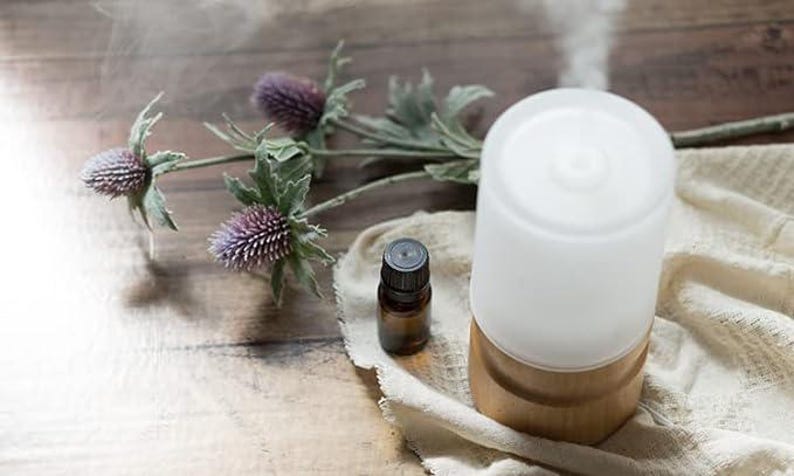 Puede incluir: Un difusor de aceites esenciales de cer&aacute;mica blanca con una base de madera se encuentra sobre un pa&ntilde;o beige con una peque&ntilde;a botella de aceite esencial de vidrio marr&oacute;n. El difusor est&aacute; emitiendo una columna de vapor.