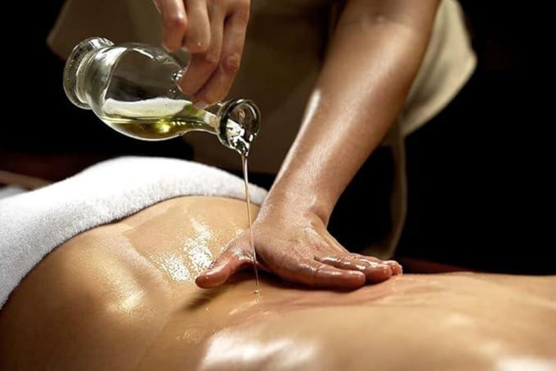 May include: A person's hand pours oil from a clear glass bottle onto a person's back during a massage.