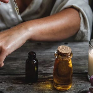 May include: A person applying essential oil to their arm with a dropper. Two amber glass bottles of essential oil and a lit candle with purple flowers are on a wooden table.