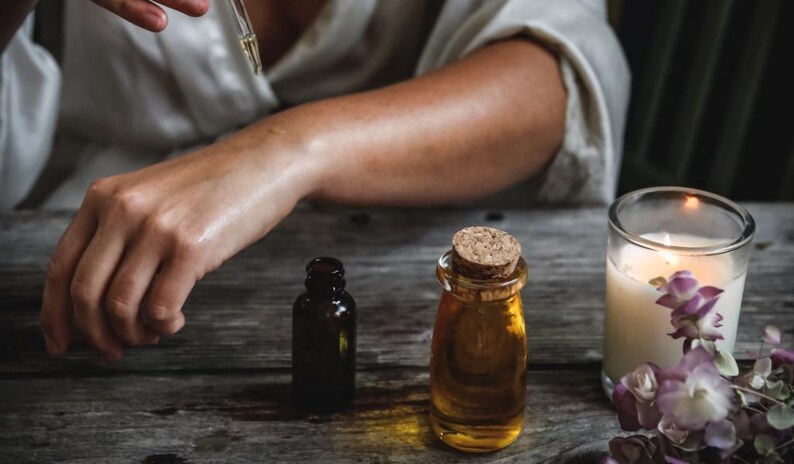 May include: A person applying essential oil to their arm with a dropper. Two amber glass bottles of essential oil and a lit candle with purple flowers are on a wooden table.