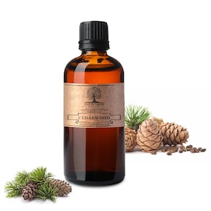 Puede incluir: Una botella de vidrio marrón con una tapa negra llena de aceite esencial de madera de cedro. La botella tiene una etiqueta con el texto "Nature's Note Organics 100% Pure Aromatherapy Essential Oil Cedarwood naturesnotenorganics.com". La botella está sobre una superficie blanca con piñas y agujas.