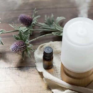 May include: A white ceramic essential oil diffuser with a wooden base sits on a beige cloth with a small brown glass bottle of essential oil. The diffuser is emitting a plume of steam.