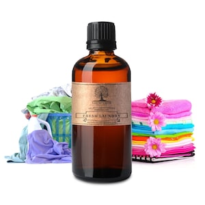 May include: A brown glass bottle with a black cap and a brown paper label. The label says "Nature's Note 100% Pure Aromatherapy Essential Oil Fresh Laundry naturesnotesorganics.com". The bottle is sitting on a white background with a pile of colorful laundry in the background.