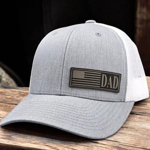 May include: Gray trucker hat with a white mesh back and a curved brim. The hat features a rectangular patch with an American flag design and the word "DAD" in a bold font. The hat is sitting on a wooden surface.