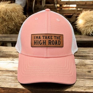 May include: A pink and white trucker hat with a brown leather patch that says "I'm a take the high road".