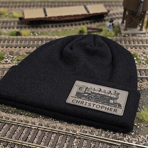 May include: A black knit beanie featuring a rectangular tan patch on the front. The patch displays a detailed illustration of a steam locomotive train and the name CHRISTOPHER printed in bold, capitalized black letters below the train graphic.
