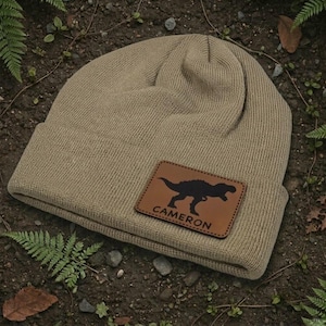 May include: A tan knit beanie with a folded cuff featuring a rectangular brown leather patch. The patch is embossed with a black silhouette of a dinosaur and the name CAMERON in capital letters, centered on the front of the cuff.