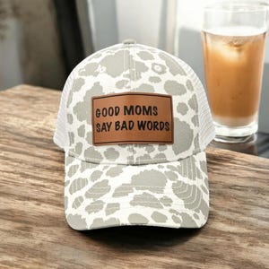 May include: A white and gray trucker hat with a brown leather patch that says "Good Moms Say Bad Words". The hat has a mesh back and a white and gray leopard print pattern.