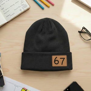 May include: A black knit beanie with a brown rectangular patch displaying the number "67". The beanie is folded at the brim and sits on a light wooden surface. Other items on the surface include a notebook, pens, glasses, and a smartphone.