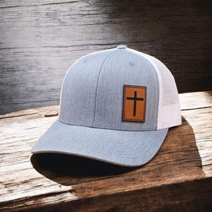 May include: A gray baseball cap with a white mesh back and a brown leather patch featuring a black cross. The cap has a curved brim and a button on top. The hat is sitting on a wooden surface.