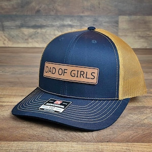May include: A navy blue and gold trucker hat with a brown leather patch that reads "Dad of Girls".