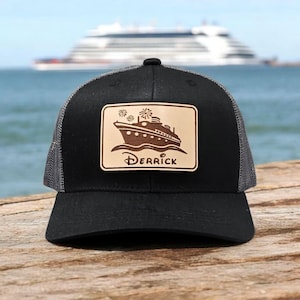 May include: A black baseball cap with a mesh back and a rectangular tan leather patch on the front. The patch features a dark brown graphic of a cruise ship with fireworks above it and the name DERRICK written in a stylized font below.