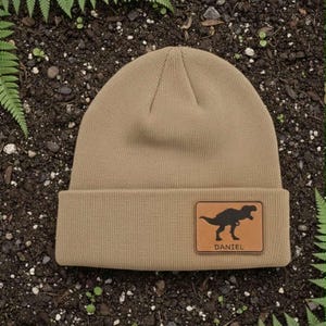 May include: A tan beanie with a rectangular leather patch featuring a black dinosaur silhouette and the name "DANIEL" in white. The beanie is made of a soft, knitted material.