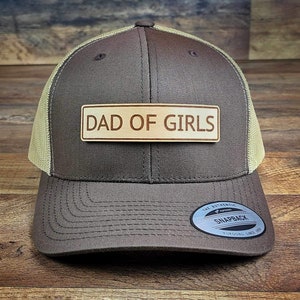 May include: A brown and tan trucker hat with a brown leather patch that says "Dad of Girls". The hat has a mesh back and a snap closure.