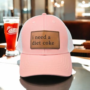 May include: Pink trucker hat with a white mesh back and a brown leather patch that says "i need a diet coke".