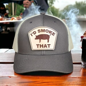 May include: A gray and beige trucker hat with a brown wooden patch that says "I'd Smoke That" and features a silhouette of a pig.