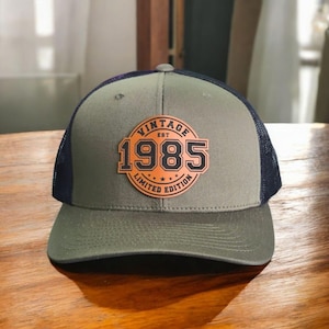 May include: A green and black trucker hat with a brown leather patch that says "Vintage Est 1985 Limited Edition".