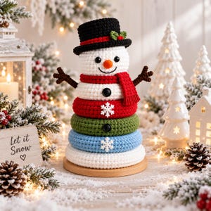 May include: A crocheted snowman stacking toy featuring a black top hat with a green leaf and red berry accent, a white head with an orange nose, and a red scarf. The body consists of stacked rings in red, green, blue, and white with snowflake details.