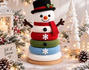 Peut inclure: Un jouet à empiler en forme de bonhomme de neige au crochet, doté d'un haut-de-forme noir avec une feuille verte et une baie rouge, une tête blanche avec un nez orange et une écharpe rouge. Le corps se compose d'anneaux empilés rouges, verts, bleus et blancs.