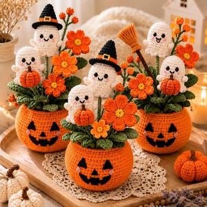 May include: Three crocheted arrangements in orange pumpkin-shaped pots with jack-o'-lantern faces. Each features white ghost figures, some wearing black witch hats, orange flowers, green leaves, and a small brown broomstick, with extra pumpkins.
