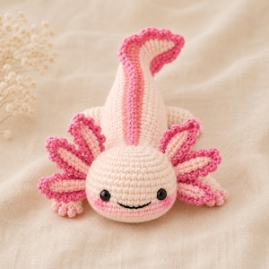 Peut inclure: Une peluche axolotl crochetée présentant un corps rose pâle avec des branchies externes rose vif et une nageoire dorsale assortie. La créature possède de petits yeux en perles noires et un sourire cousu simple, réalisé en mailles texturées.