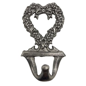 May include: A silver-toned metal wall hook featuring a heart-shaped top adorned with embossed floral patterns and a central bow detail. The hook has a sturdy base with a curved, double-pronged design, providing a functional element for hanging items.