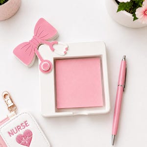 Op de afbeelding: Een witte vierkante notitieblokhouder met een roze notitieblok, een roze pen en een roze strikvormige stethoscoop. Een witte en roze verpleegsterbadgehouder en een kleine potplant zijn ook zichtbaar.