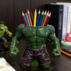 May include: A green ceramic desk organizer shaped like the Hulk's torso, holding colorful pencils. The figure has defined muscles and purple shorts. A small Hulk figurine is visible in the background. The pencils are various colors, including red, yellow, and blue.