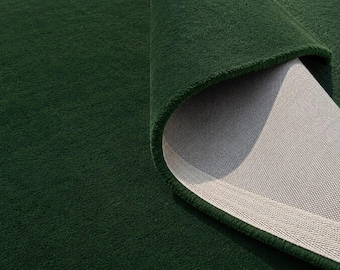 May include: A deep forest green area rug with a dense, short-pile texture. One corner of the rug is folded over to reveal the light beige, woven fabric backing and a reinforced, herringbone-patterned edge binding along the underside perimeter.