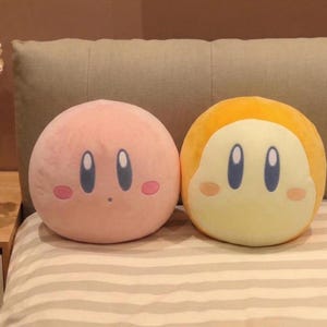Puede incluir: Dos almohadas redondas de felpa. Una es rosa con ojos azules y mejillas rosadas. La otra es amarilla y blanca con ojos azules y mejillas rosadas. Las almohadas están sobre una cama con un estampado de rayas.