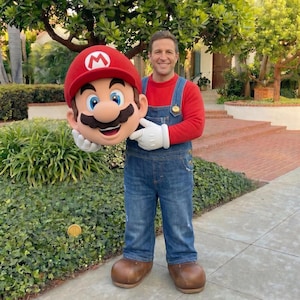 Puede incluir: Una persona con una camisa roja y un mono vaquero azul, sosteniendo una gran cabeza de Mario. La cabeza de Mario es roja con una "M" blanca en el sombrero, ojos azules y un gran bigote. La persona lleva guantes blancos y zapatos marrones.