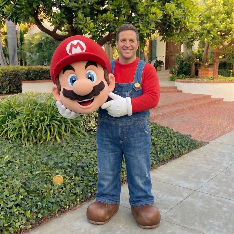 Puede incluir: Una persona con una camisa roja y un mono vaquero azul, sosteniendo una gran cabeza de Mario. La cabeza de Mario es roja con una "M" blanca en el sombrero, ojos azules y un gran bigote. La persona lleva guantes blancos y zapatos marrones.
