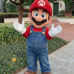 Puede incluir: Una persona con un disfraz de Mario, con un gorro rojo con una "M" blanca, una camisa roja, un peto vaquero azul, guantes blancos y zapatos marrones. El personaje est&aacute; de pie en una acera con un seto verde y una casa al fondo.