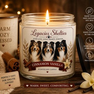 Op de afbeelding: Een brandende Cinnamon Vanilla geurkaars in een glazen pot, met portretten van Sheltie honden. Het etiket luidt "Legacies Shelties" en "Warm, Sweet, Comforting." Op de achtergrond staat een mok met "Warm, Cozy, Blessed".