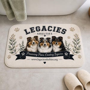 May include: A rectangular beige bath mat featuring three Sheltie dog portraits. The mat has the text "LEGACIES SHELTIES" above a banner that reads "Preserving Paws, Creating Legacies" and the website address. Decorative paw prints and foliage surround the design.