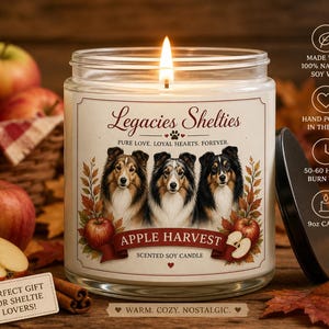 Op de afbeelding: Een brandende glazen potkaars met een zwart deksel, met een etiket met drie Sheltie-hondenportretten en de tekst "Apple Harvest". De kaars is omgeven door appels, kaneelstokjes en herfstbladeren. De kaars is 255g.