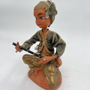 Op de afbeelding: Een vintage beeldje van een zittende figuur die fluit speelt. De figuur draagt een groene fluwelen jas met gouden rand, een bijpassende hoofdbekleding en een gouden broek. Het beeldje is gemonteerd op een perzikroze voet met een goudkleurig dier.