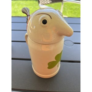 Peut inclure: Une tasse en céramique crème avec un visage d'oiseau fantaisiste sur le couvercle. La tasse présente un motif de trèfle vert sur le devant. La poignée est sur le côté. La tasse est sur une surface gris foncé.