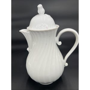 May include: White ceramic teapot with a lid and a curved handle. The teapot has a spiral design and a spout. The lid is topped with a small, decorative flower. The teapot is set against a dark background.