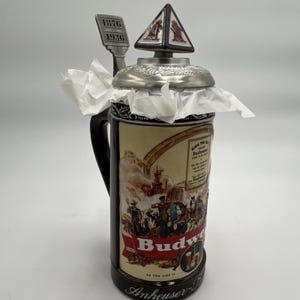 Può includere: Un boccale di birra Budweiser vintage con manico e base neri. Il boccale presenta un'illustrazione dettagliata e il logo Budweiser. Il coperchio è argentato con una parte superiore decorativa. Il boccale è probabilmente un oggetto da collezione.